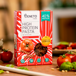 Dieses Bild zeigt einen Anwendungsfall, wie die Beneto Pasta Verpackung Zuhause in der Küche aussehen könnte. Die Faltschachtel steht auf einer Küchenplatte mit Tomaten, Pinienkernen, frischen Basilikumblättern, einem Teller mit Nudeln und einem Kochlöffel. Der Hintergrund ist verschwommen.